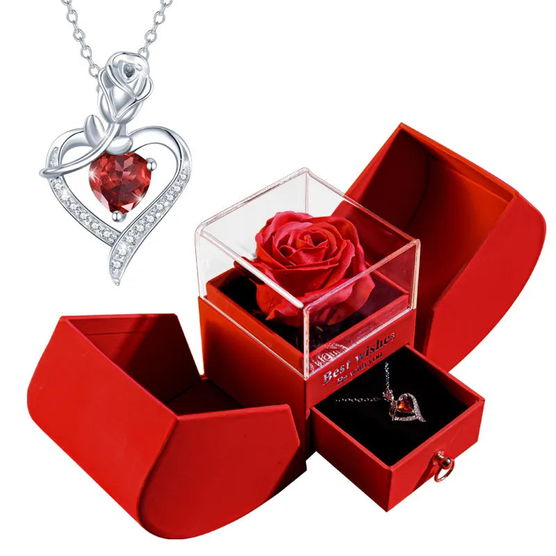A silver heart-shaped necklace with a red rose pendant and clear crystal accents, shown alongside a red gift box set. The box includes a square clear case holding a preserved red rose and a small drawer containing the necklace.